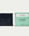 A dark charcoal soap brick next to its mint green packaging box, both embossed with the brand name 'FLAMINGO ESTATE'. A bright green circular badge in the top right corner reads 'NEW LOOK SAME FORMULA'.