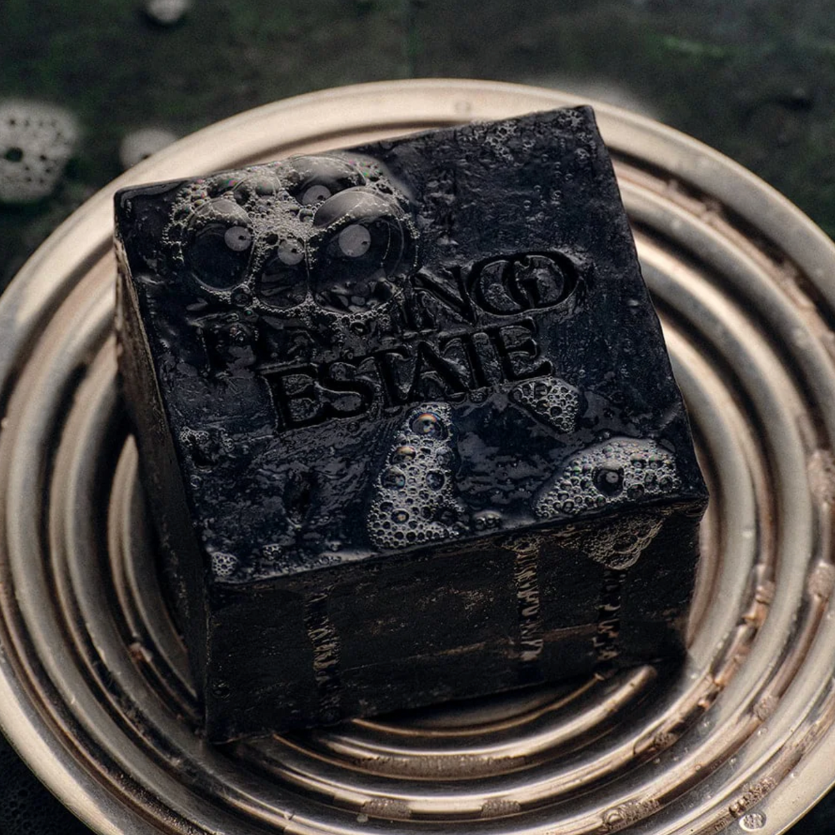 A dark, textured square bar of soap with 'FALCON ESTATE' embossed on the top, resting on a metallic circular dish against a dark, wet background.