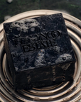 A dark, textured square bar of soap with 'FALCON ESTATE' embossed on the top, resting on a metallic circular dish against a dark, wet background.