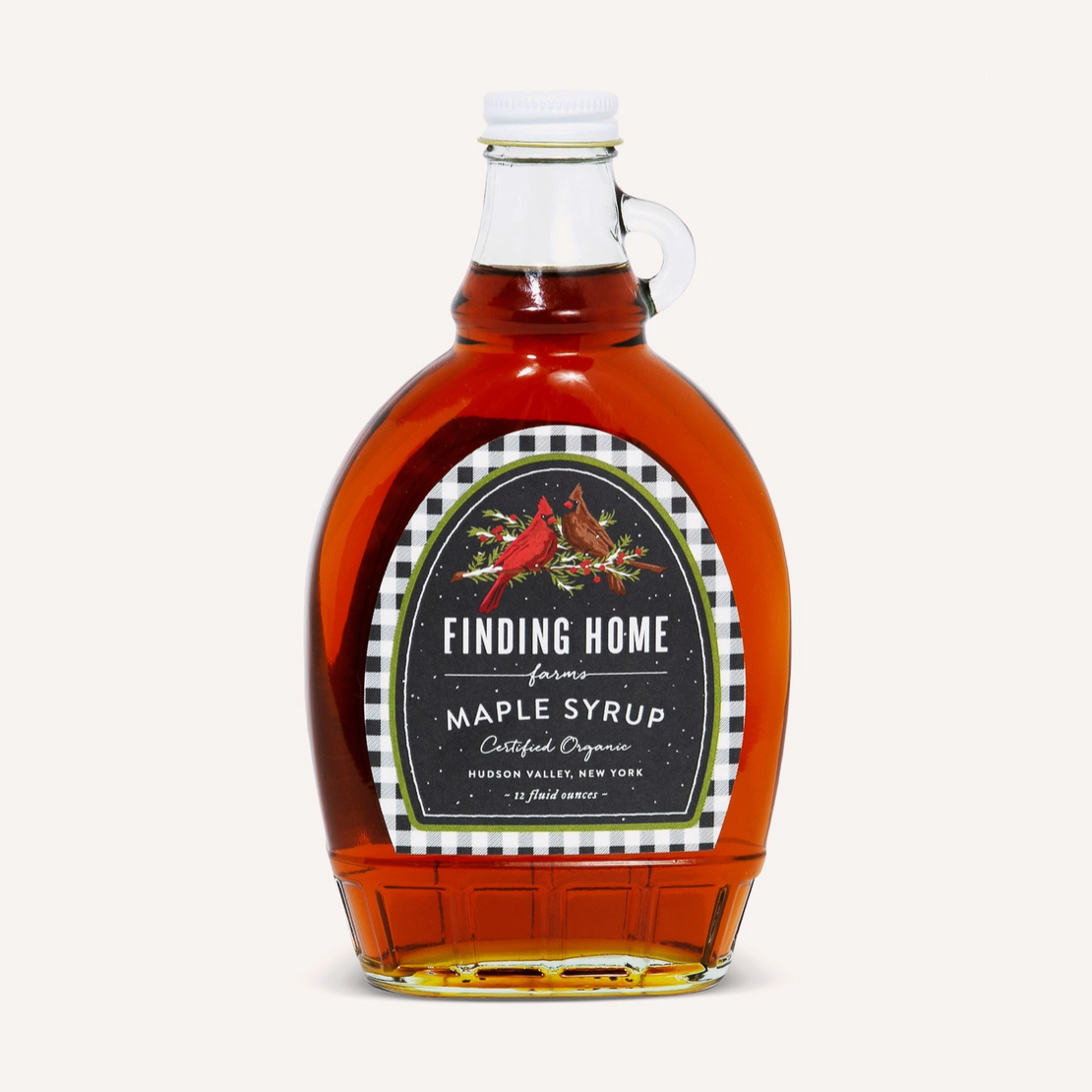 A bottle of Finding Home from Hudson Valley, New York maple syrup, filled with amber liquid, featuring a white cap and a decorative black, red, and white label, set against a solid off-white background.