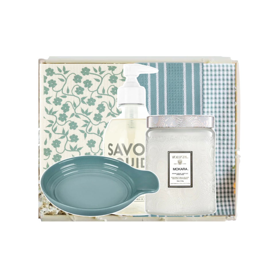 A gift set in a clear box featuring a blue-green ceramic dish, a frosted glass jar of Mokaia hand wash, a pump bottle of Savonique hand wash, and folded towels with blue and white patterns.