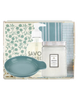 A gift set in a clear box featuring a blue-green ceramic dish, a frosted glass jar of Mokaia hand wash, a pump bottle of Savonique hand wash, and folded towels with blue and white patterns.