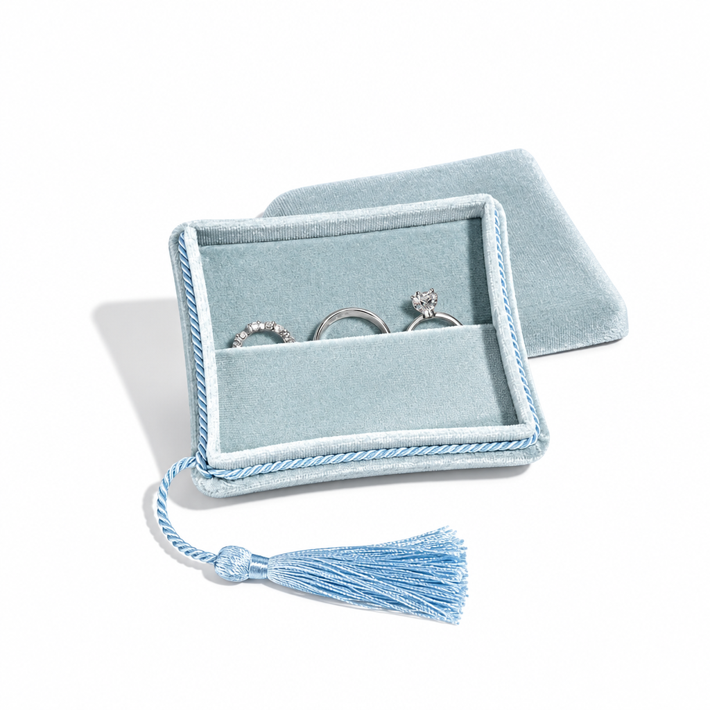 A light blue velvet ring box with a tassel, shown open with three silver rings inside on a white background.