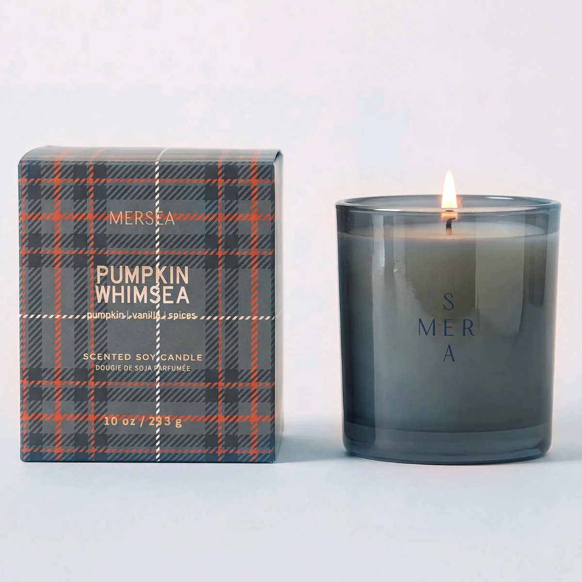 A lit Mersa 'Pumpkin Whimsea' scented soy candle in a dark gray glass jar next to its retail box featuring a gray, orange, and black plaid pattern.