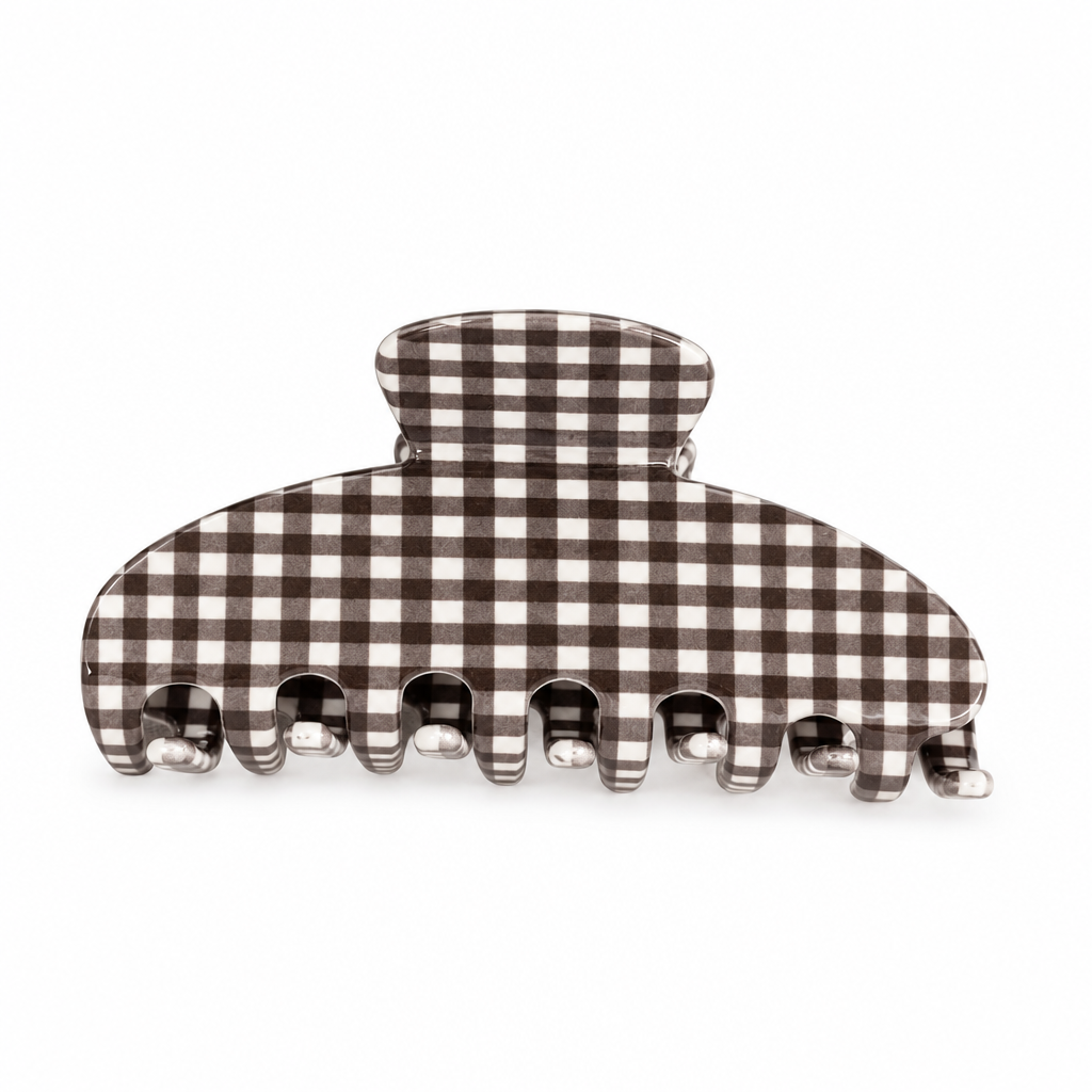 A claw hair clip with a brown and beige gingham checkered pattern, resting on a textured beige linen surface with a woven straw basket in the background.
