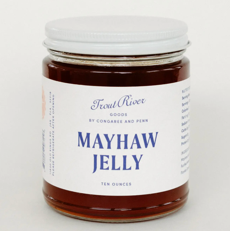 A glass jar of Trout River Goods Mayhaw Jelly, filled with dark amber jelly and featuring a white label with blue text and a white metal lid, set against a plain light background.