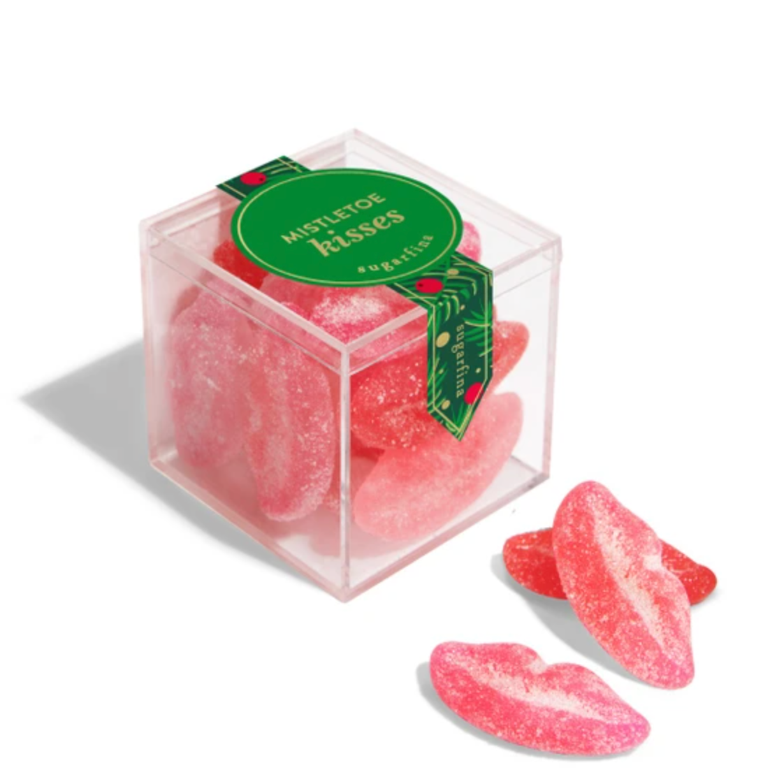 A clear square box filled with pink sugar-coated gummy candies shaped like lips, with three loose lip-shaped gummies placed in front on a white background. A green label on the box reads 'MISTLETOE kisses' and 'Kool-Aid'.