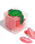 A clear square box filled with pink sugar-coated gummy candies shaped like lips, with three loose lip-shaped gummies placed in front on a white background. A green label on the box reads 'MISTLETOE kisses' and 'Kool-Aid'.