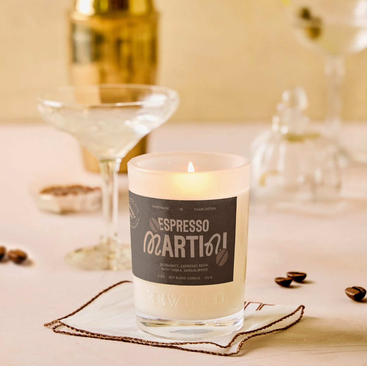 A lit scented candle in a clear glass jar sits on a napkin on a tabletop, with coffee beans scattered around it. The label reads 'Nespresso Marti 01'.