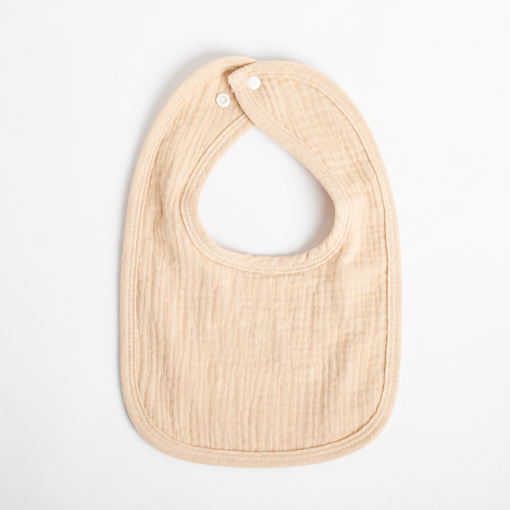 A personalized baby bib in a soft, textured muslin fabric in a light beige color, featuring the name 'Blake' embroidered in white on the front. The bib has a rounded bottom, a curved neck opening, and two white snap buttons at the top, shown flat on a clean white background.
