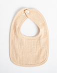 A personalized baby bib in a soft, textured muslin fabric in a light beige color, featuring the name 'Blake' embroidered in white on the front. The bib has a rounded bottom, a curved neck opening, and two white snap buttons at the top, shown flat on a clean white background.