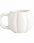 A white ceramic mug with a ribbed pumpkin-like design sits on a white marble tray on a kitchen counter, with apples and a blurred plant in the background.