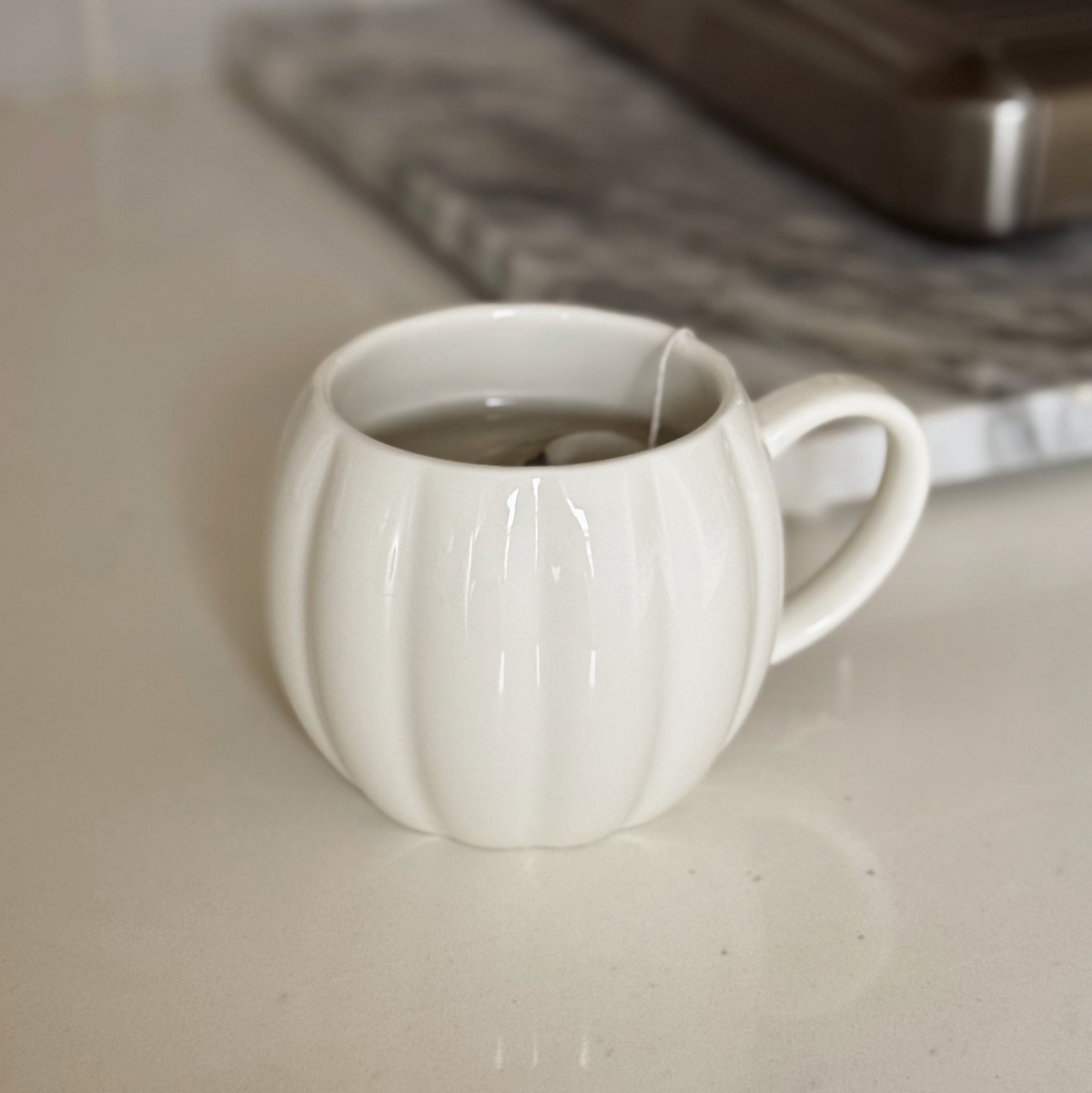 A white ceramic mug with a ribbed pumpkin-like design, filled with tea and a tea bag, sitting on a light countertop with a blurred kitchen background.