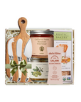 A gourmet gift basket set containing a wooden cutting board with three knives, a jar of California Native Mountain Wild Turkey Honey, a jar of Rustic Bakeri Rosemary and Olive Oil, a pack of Rustic Ranch Salami, and a pack of Rustic Ranch Anch & Hickory Bread, all arranged in a white gift box with a green ribbon.