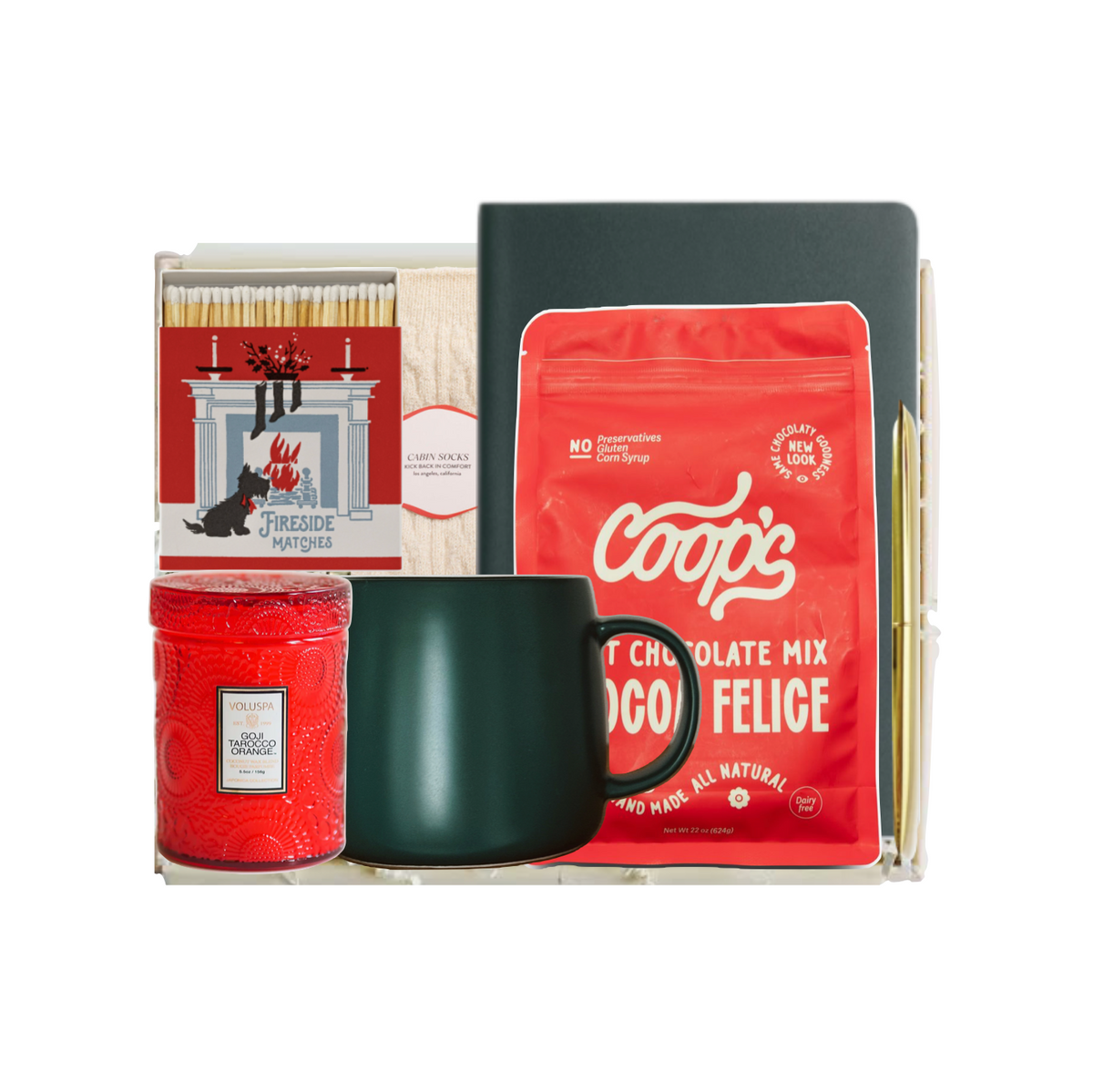 A curated gift basket set in a clear acrylic tray, featuring a red bag of Coop's Hot Chocolate Mix, a dark green ceramic mug, a red candle, a box of matches, and a black notebook, isolated on a white background.