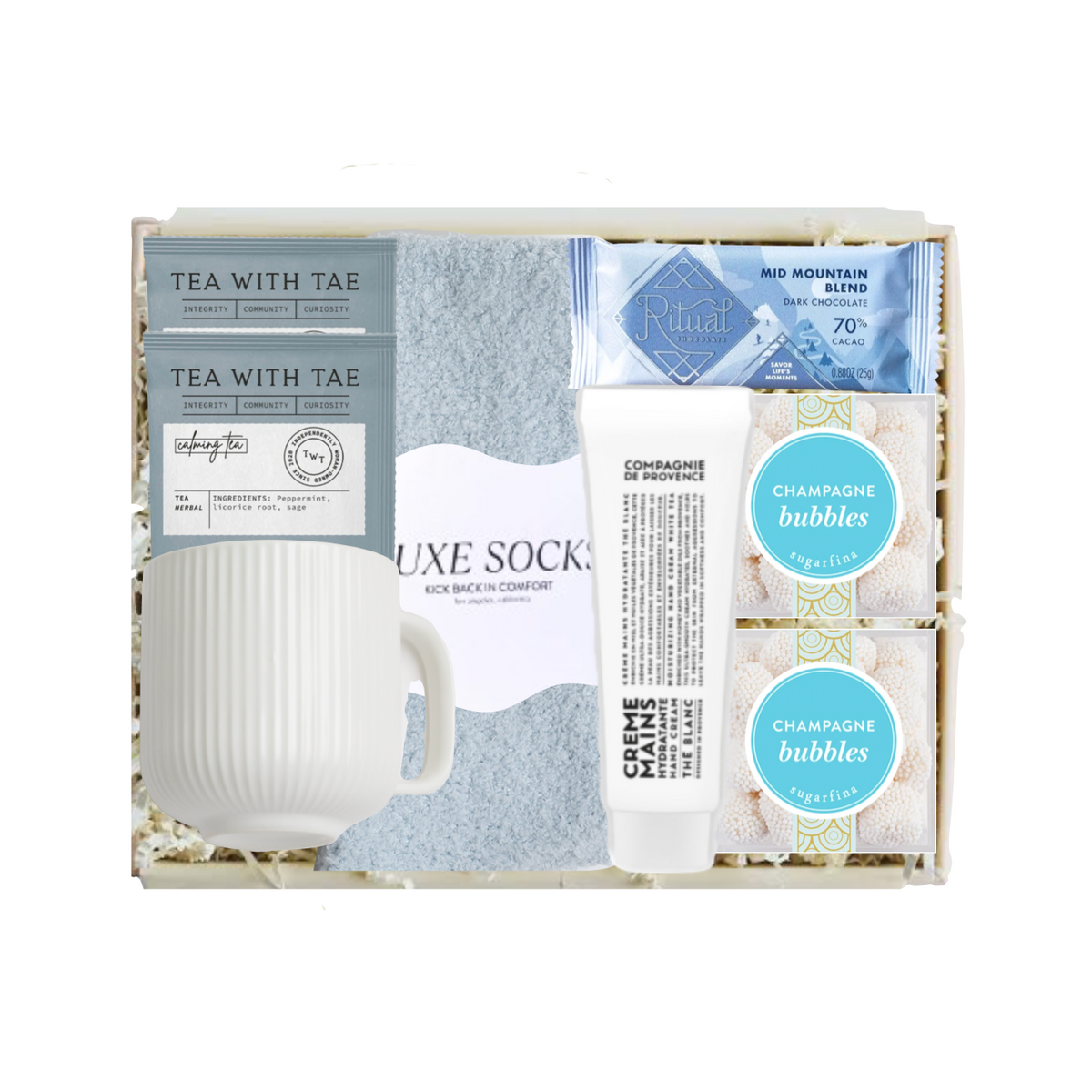 A curated gift box set containing a white ribbed ceramic mug, a light blue textured sock, a tube of Crème Brûlée spread, two round bath bombs labeled 'Champagne Bubbles', and two tea bags from 'Tea With Tae'.