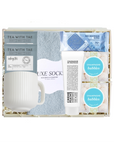 A curated gift box set containing a white ribbed ceramic mug, a light blue textured sock, a tube of Crème Brûlée spread, two round bath bombs labeled 'Champagne Bubbles', and two tea bags from 'Tea With Tae'.