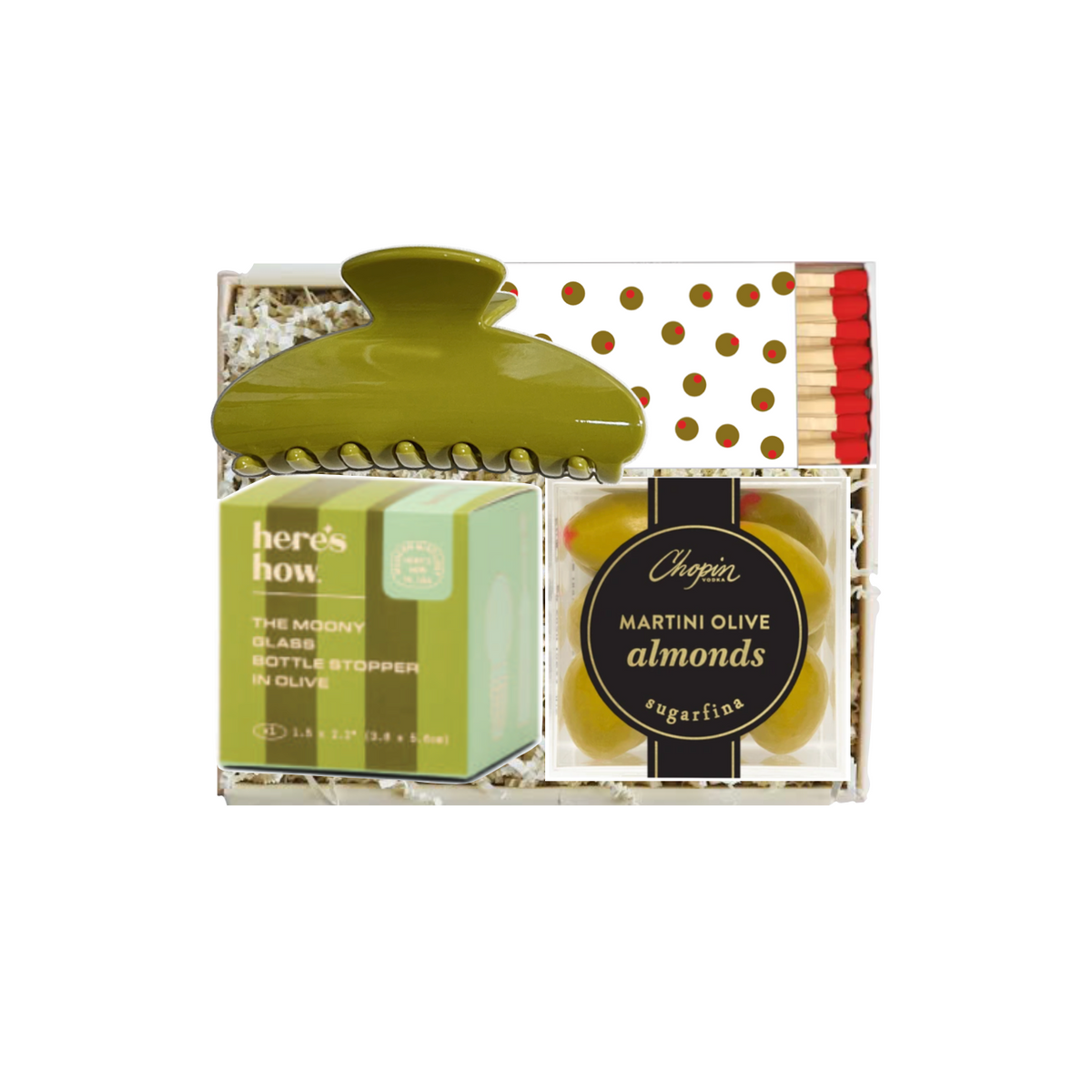 A gift set containing a green hair claw clip, a box of Here's How soap, and a bag of Chogokin Martini olives and almonds, arranged in a clear gift box with shredded paper and gold polka dots.