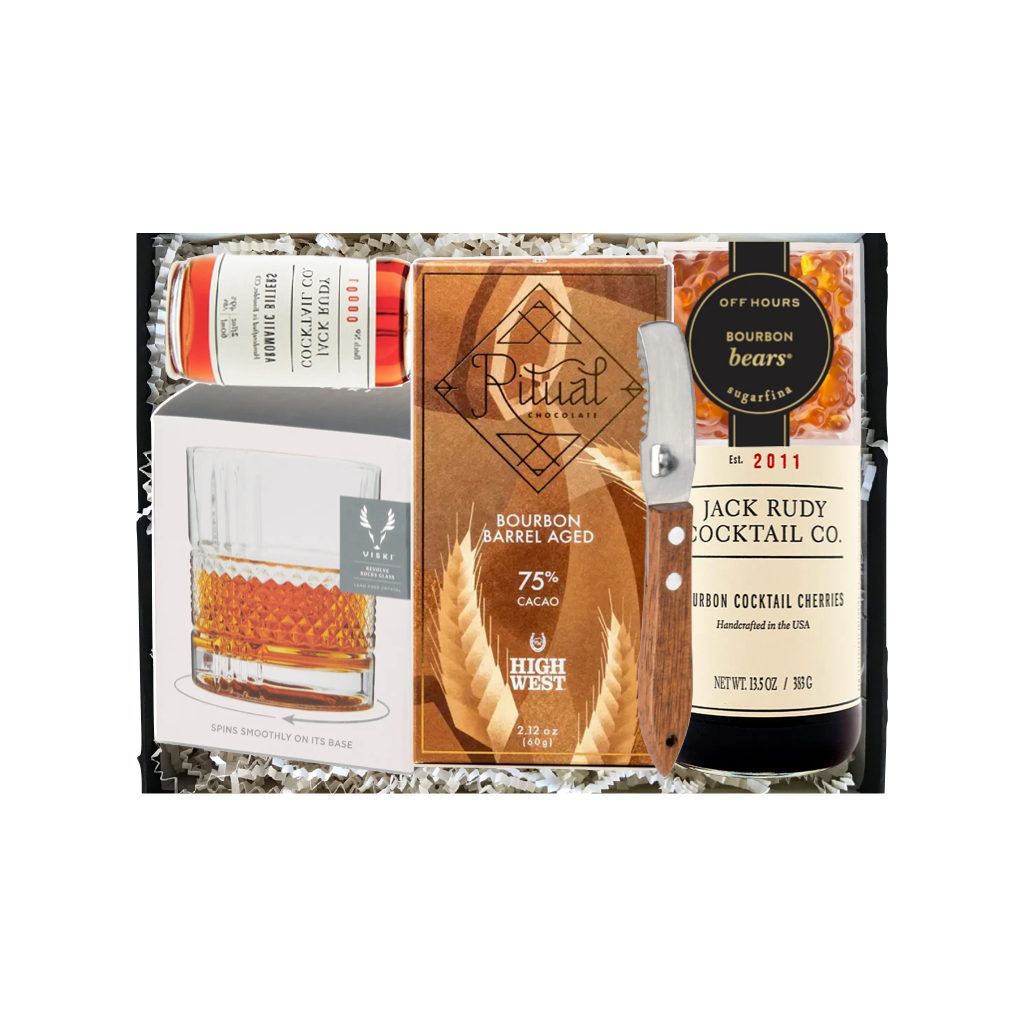 A curated whiskey gift set in a black tray filled with white paper shred, featuring a bottle of Jack Rudy Cocktail Co. Bourbon Cocktail Crèche, a Ritual 75% Aged Bourbon Barrel Aged whiskey bottle, a clear glass tumbler, and a branded box.