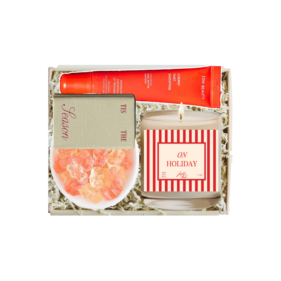 A curated gift set in a white box filled with shredded paper, featuring a red tube of hand cream, a striped candle labeled 'ON HOLIDAY', a small bowl of orange citrus peels, and a card reading 'Scented'.