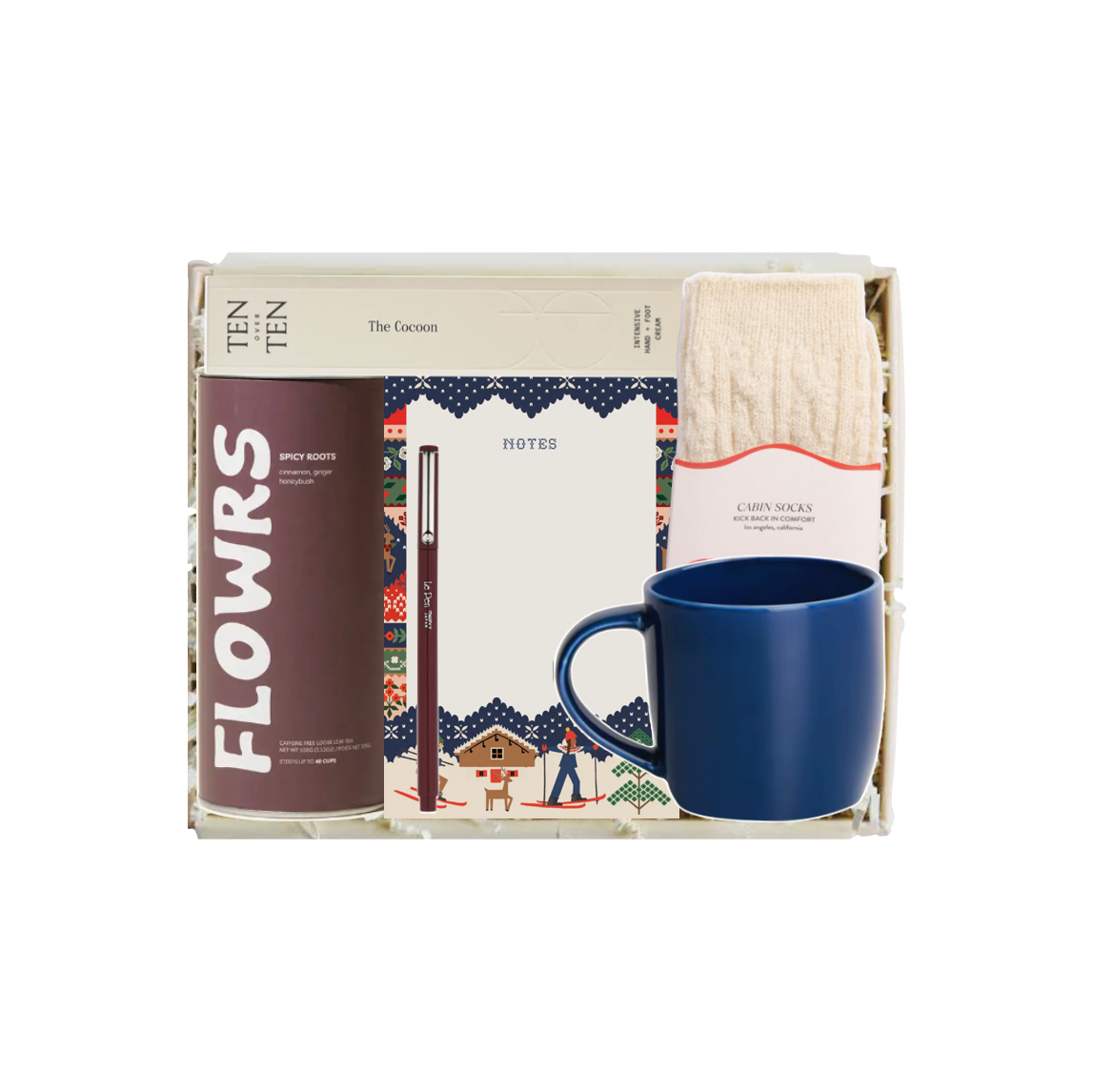A curated gift box set containing a brown and white 'FLOWRS' notebook, a white notepad with a colorful border, a cream knit sock, and a blue ceramic mug, all arranged inside a light-colored box.
