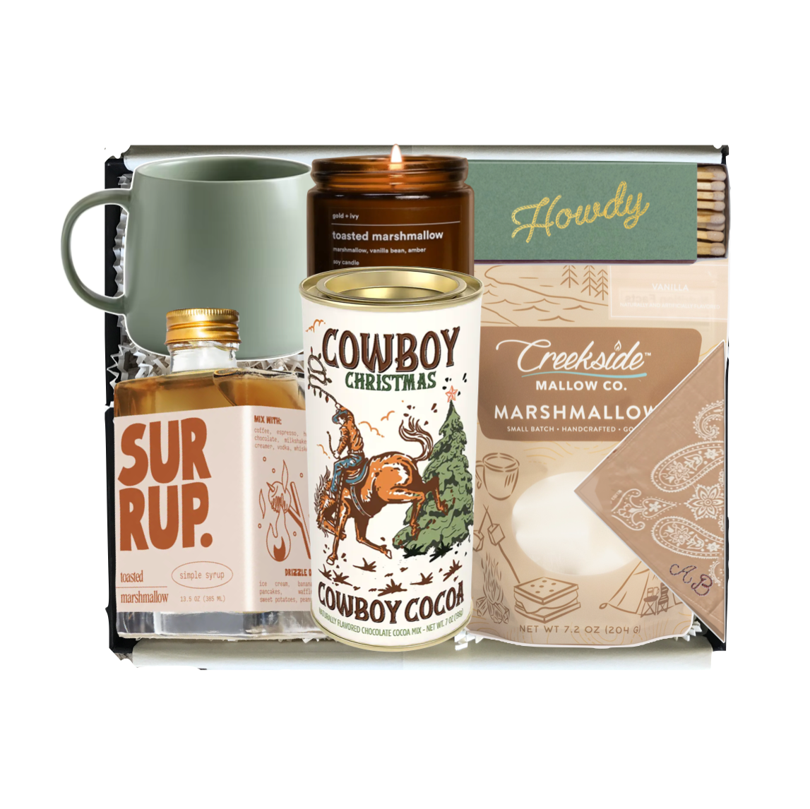 A curated gift set in a black tray featuring a sage green ceramic mug, a bottle of Surrup. handcrafted spirit, a lit candle from Cowboy Christmas, a bag of Creeside's Marshmallow, and a pack of Howdy matchsticks.