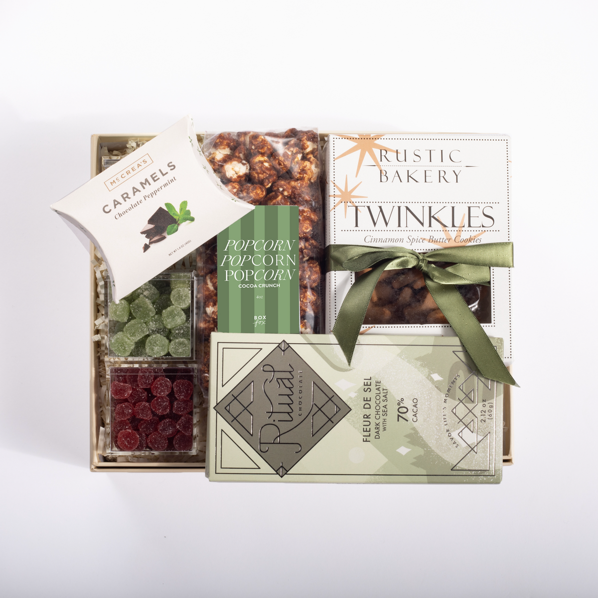 A gourmet gift box filled with assorted snacks and treats, including Rustic Bakery Twinkles, Rival chocolate, caramel corn, gummy candies, dried cranberries, and a package of popcorn, arranged on a white background.