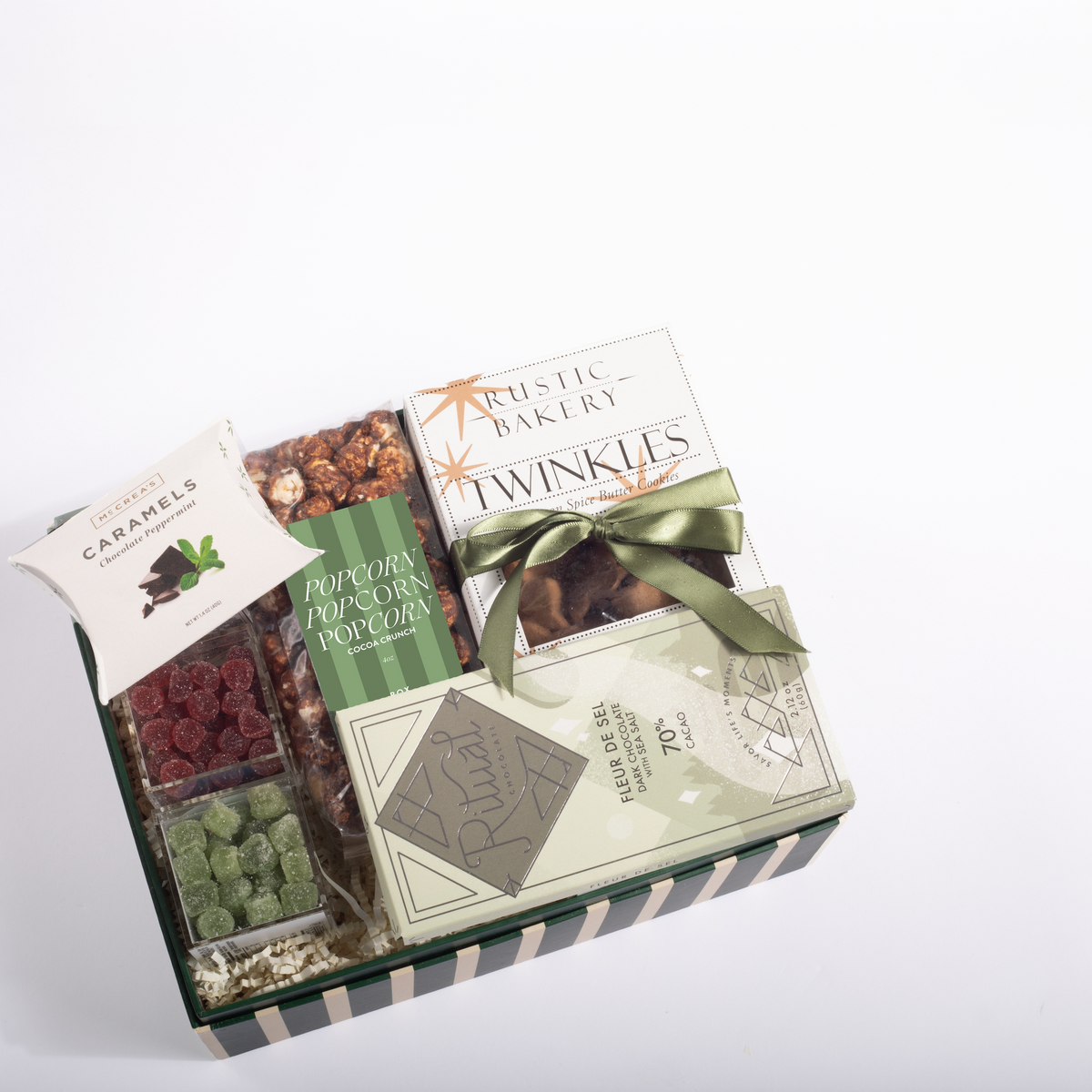 A gourmet gift box from Rustic Bakery featuring assorted chocolates, caramel candies, green pistachio-covered chocolates, and popcorn, arranged in a striped green and white box with a green ribbon.