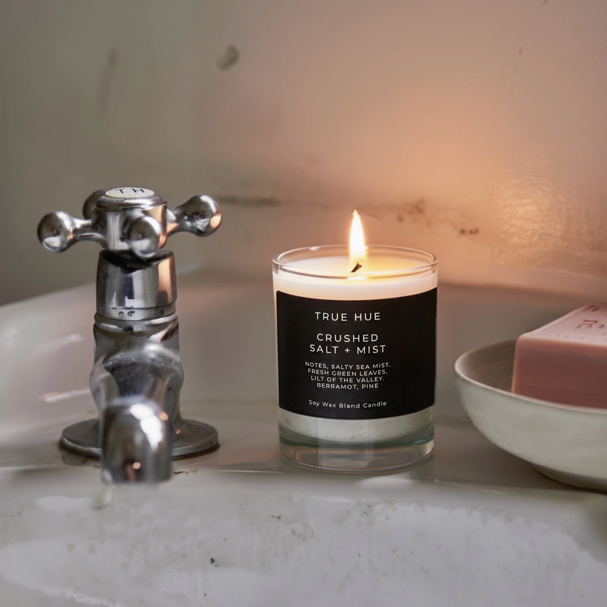 A lit True Hue 'Crushed Salt + Mist' scented candle in a clear glass jar with a black label, placed on a bathroom sink next to a chrome faucet and a pink bar of soap on a white dish.