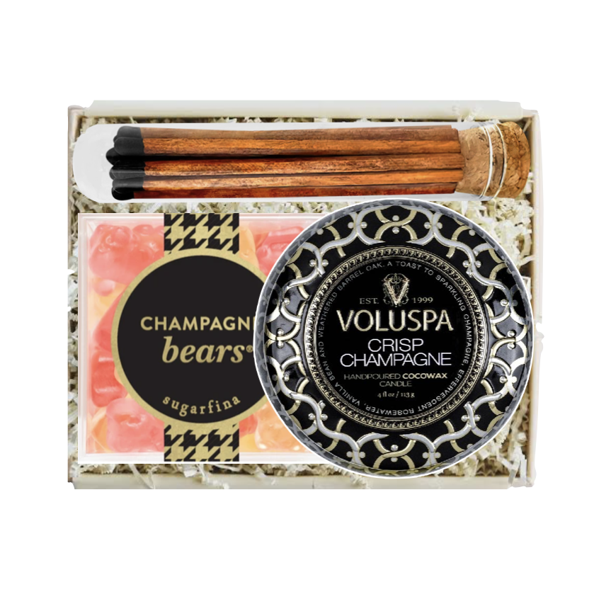 A luxury gift set in a white box containing a round tin of Voluspa Crisp Champagne handcrafted cocoa wax, a bar of champagne bears sugarfina soap, and a wooden hair stick with a black and white patterned top.