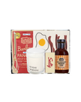 A breakfast-themed gift set from The Breakfast Club, featuring a boxed Pancake Batter mix, a 'Sunday Morning' scented candle, a bottle of Sunday Breakfast Syrup, and a small bag of bacon bits, arranged against a plain white background.
