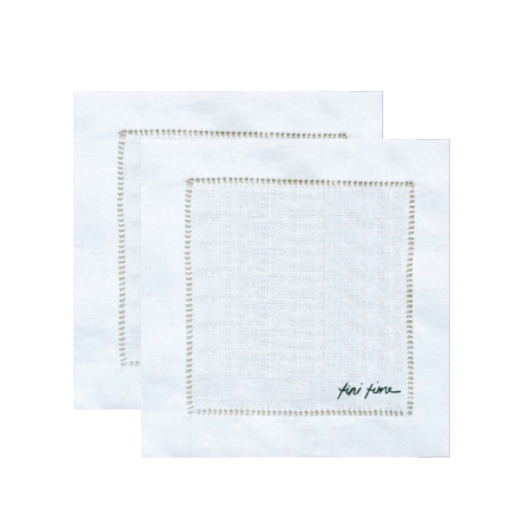 Two white linen napkins with delicate gold embroidered borders, one layered on top of the other, featuring a small black script signature on the bottom right.