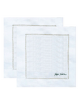 Two white linen napkins with delicate gold embroidered borders, one layered on top of the other, featuring a small black script signature on the bottom right.
