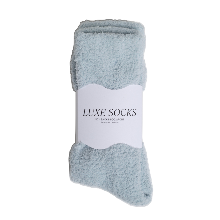 A pair of light blue fuzzy socks bundled together with a white paper label that reads 'LUXE SOCKS'.