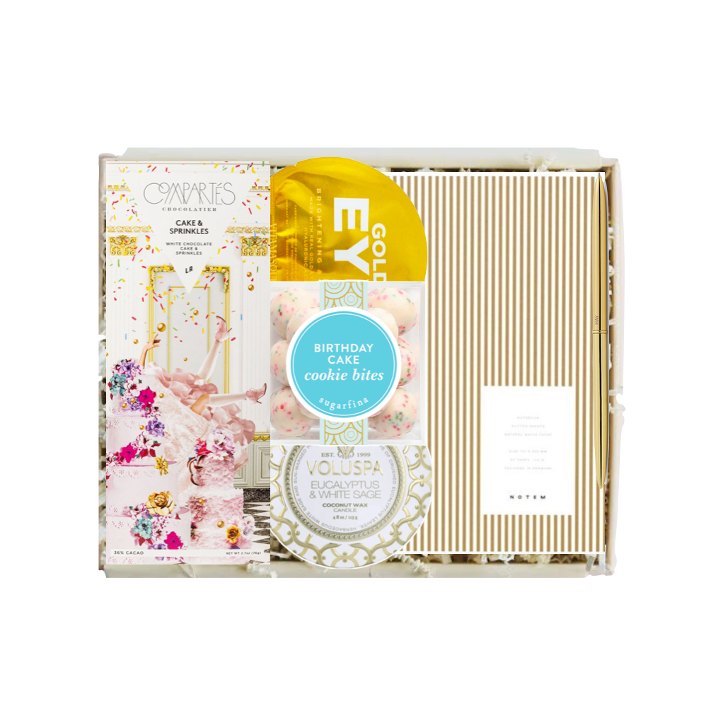 A curated gift box set from The Gift Shop featuring a 'Couples' candle, 'Eyebrow' eyelash serum, 'Voluspa' face mask, and 'Birthday Cake' cookie bites, arranged neatly inside a clear plastic box with a striped gold and white lid.