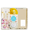 A curated gift box set from The Gift Shop featuring a 'Couples' candle, 'Eyebrow' eyelash serum, 'Voluspa' face mask, and 'Birthday Cake' cookie bites, arranged neatly inside a clear plastic box with a striped gold and white lid.