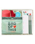 A curated gift box set containing a mint green 'Bag of Tags' pouch, a red cylindrical container, a red spiral notebook, a blue circular sticker, and a white 'List Maker' notepad with green pens, all arranged inside a clear plastic gift box on a white background.