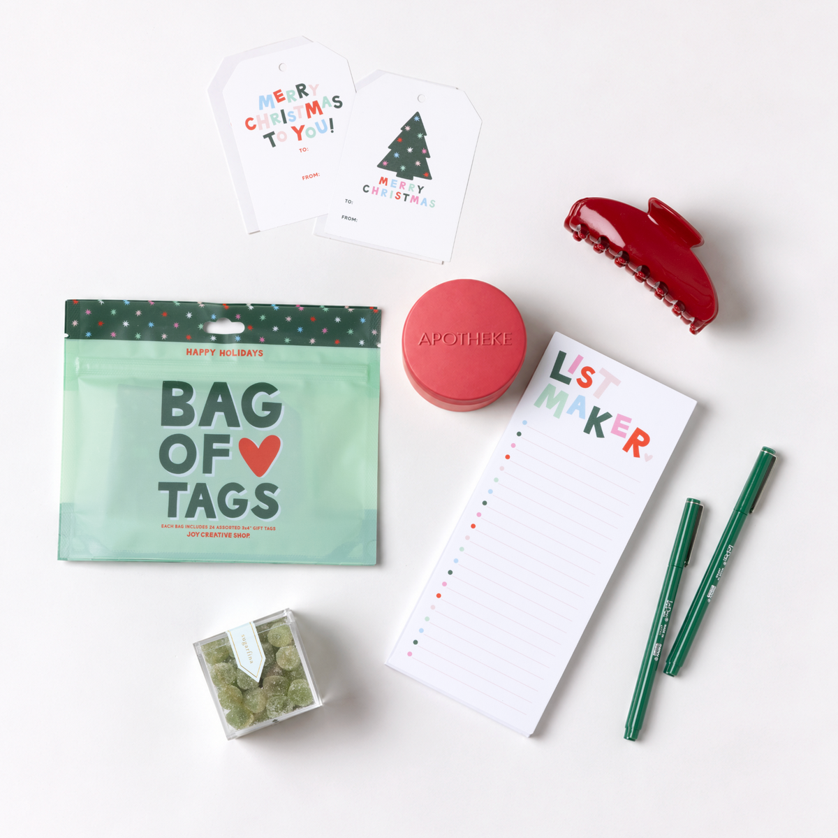 A styled flat-lay of holiday gift items on a white background, including a mint green 'Bag of Tags' pouch, a red hair claw clip, a pink round cosmetic container labeled 'APOTHEKE', a white notepad with 'List Maker' text, two green pens, and small green packaged accessories.