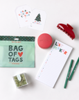 A styled flat-lay of holiday gift items on a white background, including a mint green 'Bag of Tags' pouch, a red hair claw clip, a pink round cosmetic container labeled 'APOTHEKE', a white notepad with 'List Maker' text, two green pens, and small green packaged accessories.