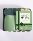 A mint green ceramic mug sits inside an open gift box with a matching green tea packet labeled 'MINTY WHITE' and a dark green scrubber, photographed on a clean white background.
