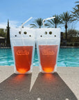 Two transparent plastic drink pouches standing upright next to a pool of water. Each pouch is holding a pink colored liquid. Water and palm trees are in the background as the pouches sit on a cement poolside. The left pouch says "Babe" in pink cursive text and the right pouch says "Bride" in white cursive text.
