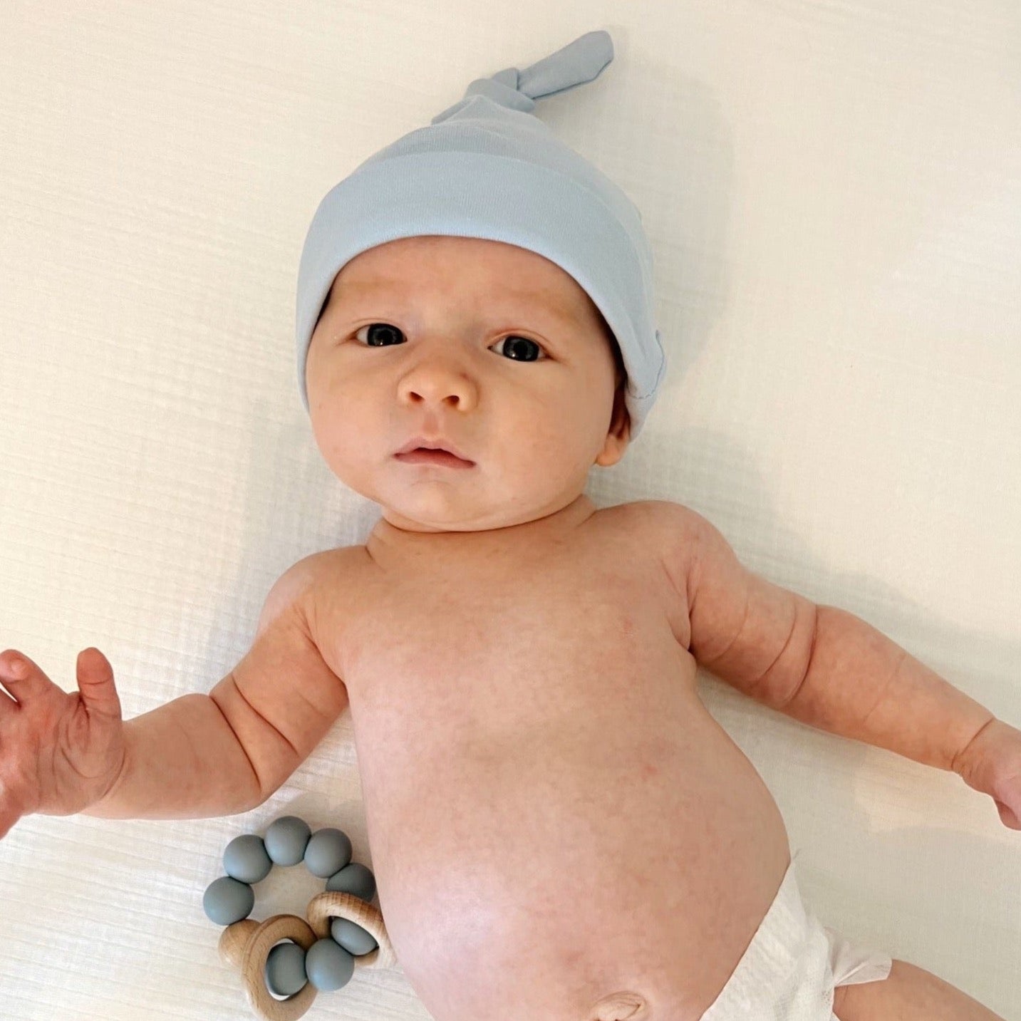 A baby wearing a blue topknot cotton hat while laying on a white muslin crib sheet.