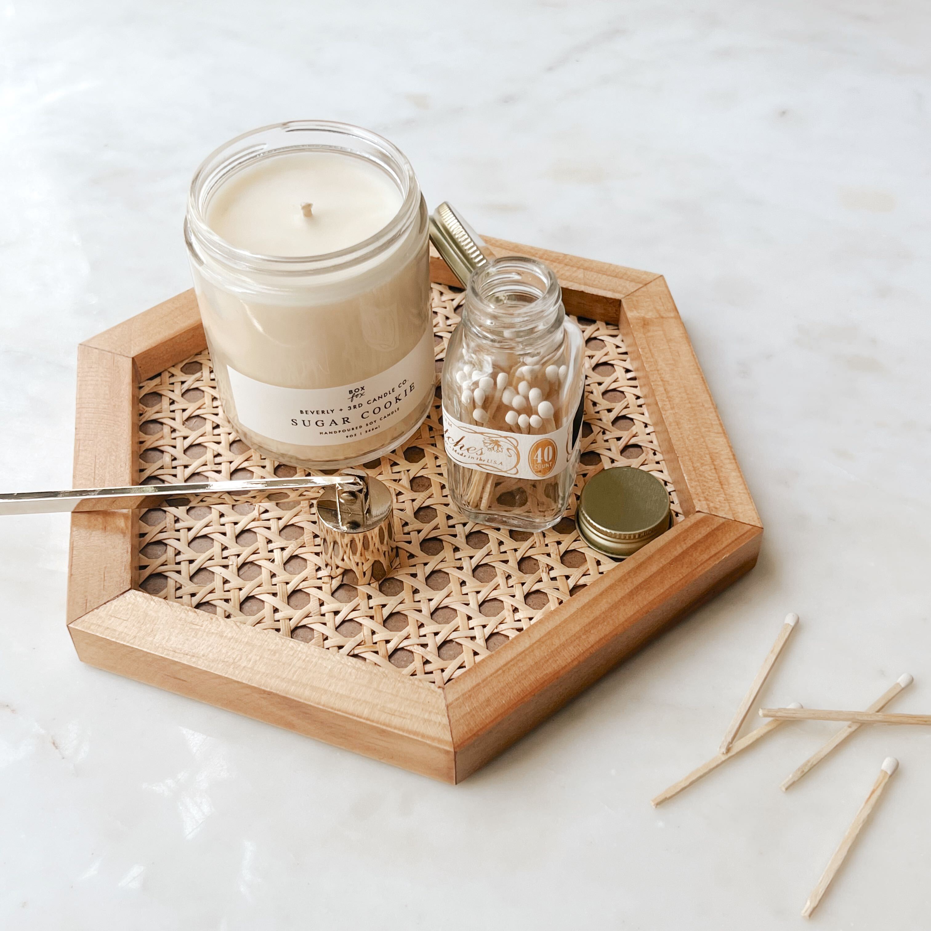 A hexagon natural wood rattan cane tray holding a sugar cookie candle, white matches, and a gold candle snuffer.
