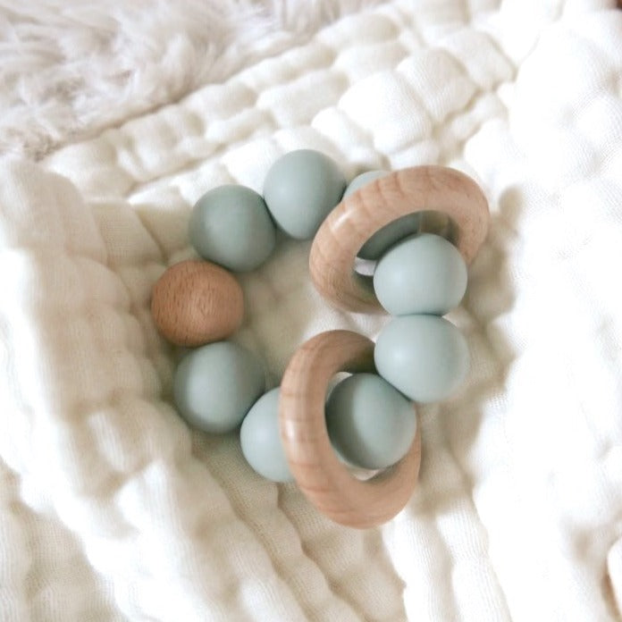 A sage green baby teether on a white muslin crib sheet.