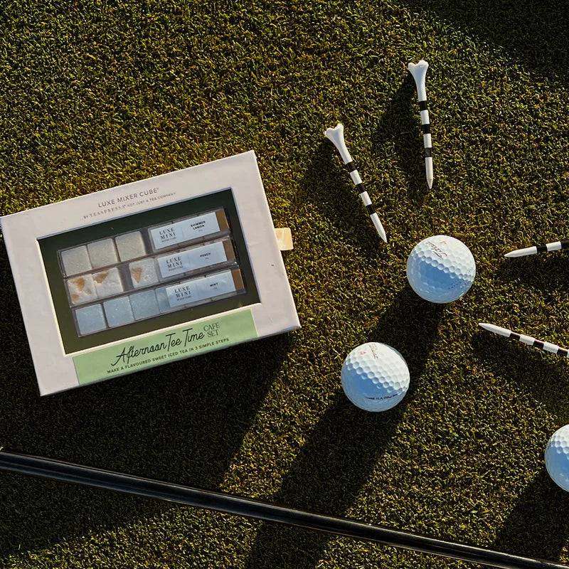 A golf-themed gift set featuring a white box labeled 'The Rich Club' and 'Adrianne's Irons' alongside several white golf balls, black-and-white striped tees, and metal golf clubs arranged on a green grass surface.