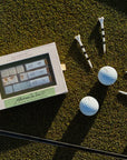 A golf-themed gift set featuring a white box labeled 'The Rich Club' and 'Adrianne's Irons' alongside several white golf balls, black-and-white striped tees, and metal golf clubs arranged on a green grass surface.