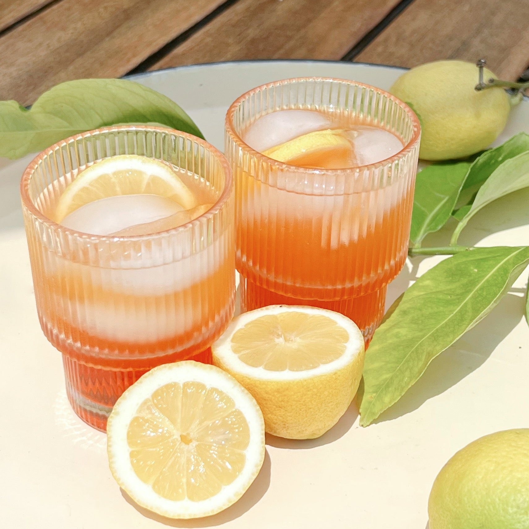A set of clear ribbed cocktail glasses sitting on a marble tray holding an orange colored drinks next to some lemon slices.