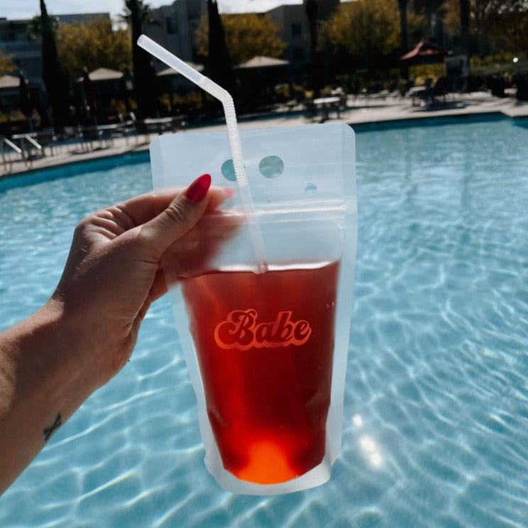 A woman&#39;s hand holding a transparent drink pouch holding pink colored liquid in front of a pool of water. On the pouch is cursive lettering spelling out the word &quot;Babe&quot; in light pink text.