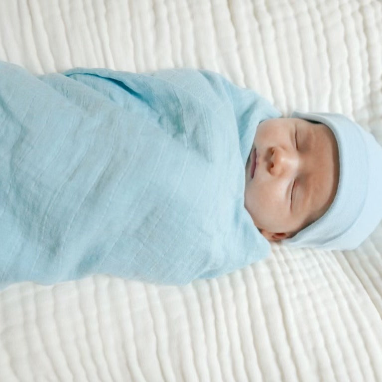 A baby wrapped in a blue snuggly cotton baby swaddle laying on a white muslin crib sheet.