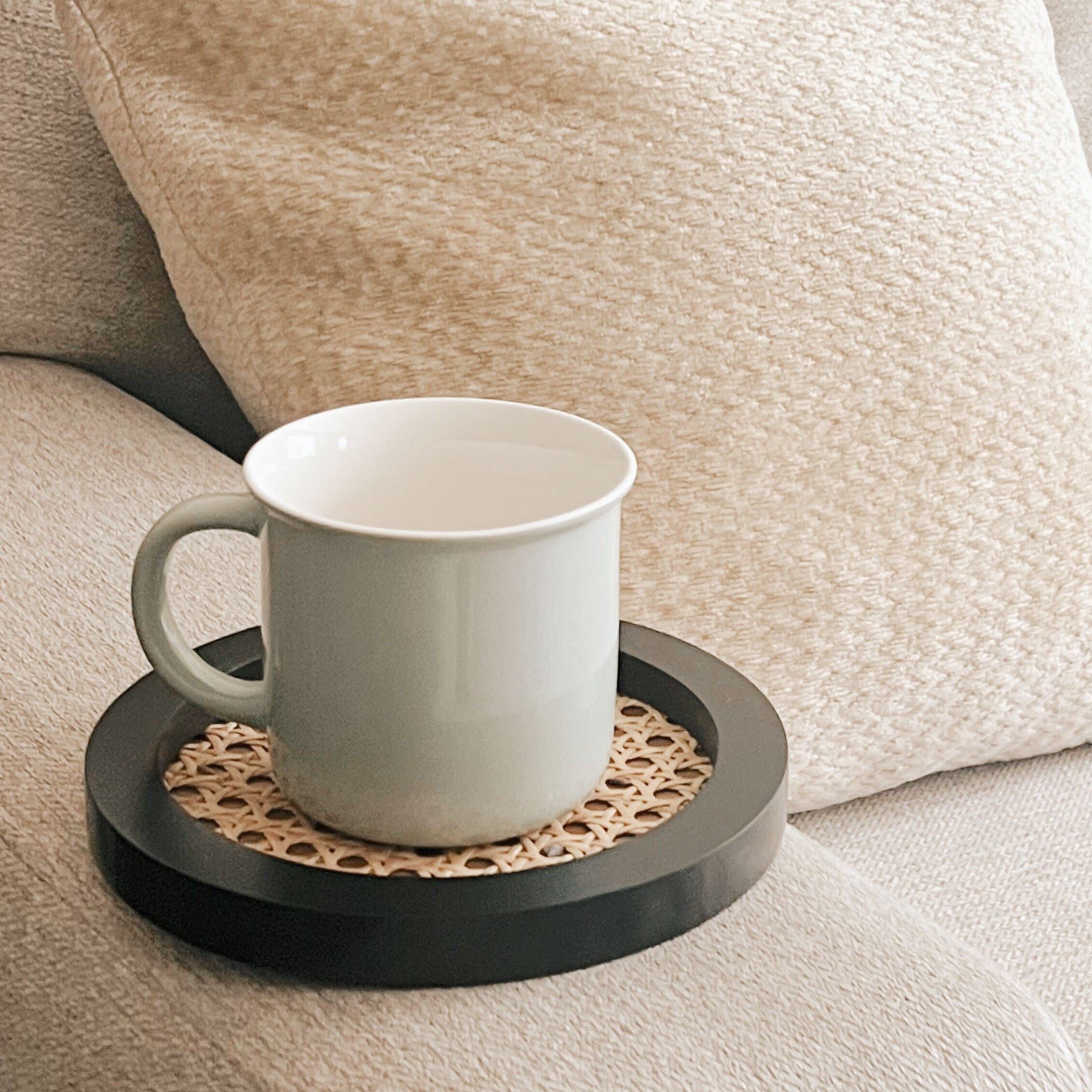 A black rattan cane tray holding a grey ceramic mug on the armrest of an oatmeal colored couch.
